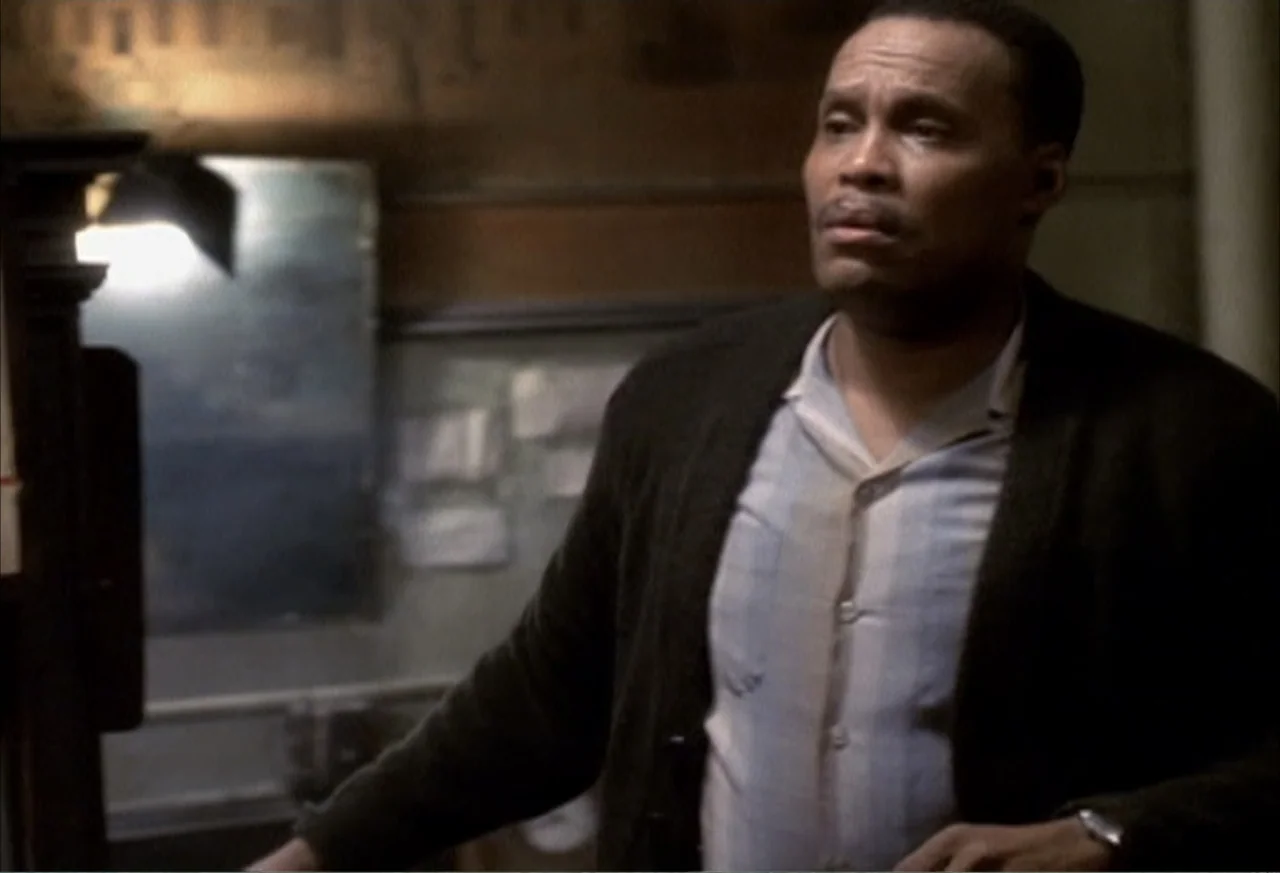 Boycott (2001) movie still: medium — An interior office space with a cluttered bulletin board and dark walls in the background. A middle-aged man…