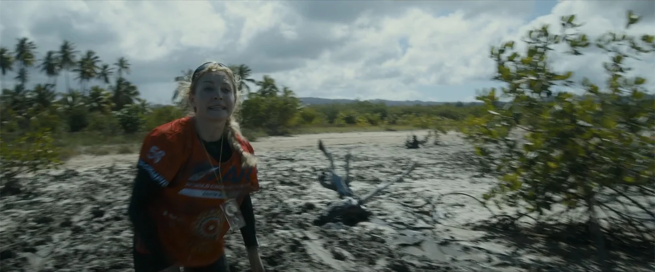 Arthur the King (2024) movie still: medium — A muddy, coastal area features low-lying vegetation and scattered driftwood under an overcast sky. A…