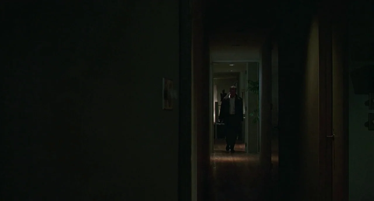 The Ice Storm (1997) movie still: wide — A dimly lit narrow residential corridor stretches away into a brightly illuminated open room. An adult male…