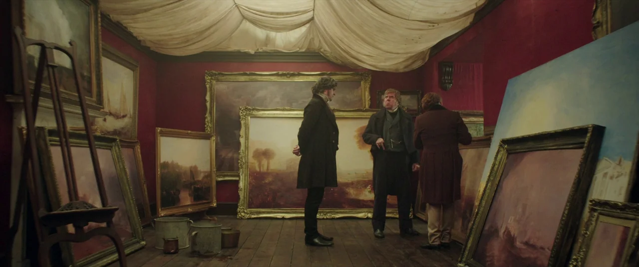 Mr. Turner (2014) movie still: wide — An 1800s art studio features red walls draped with fabric ceilings and wood flooring. Three men in period…