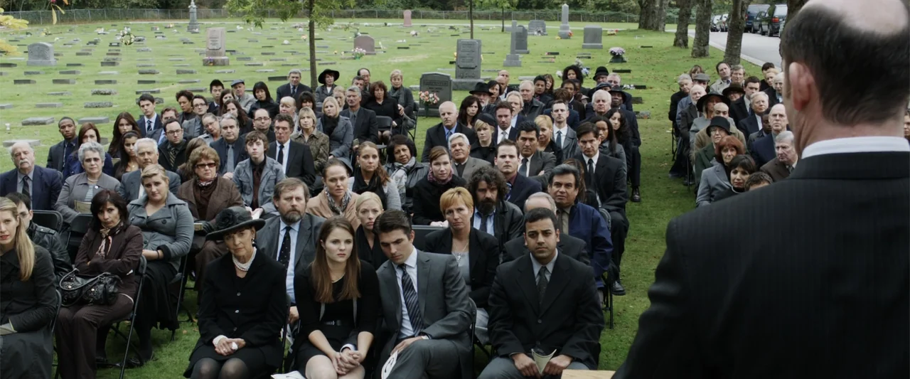 Final Destination 5 (2011) movie still: wide — An outdoor funeral service features rows of people seated in a grassy cemetery field. A man in a dark…