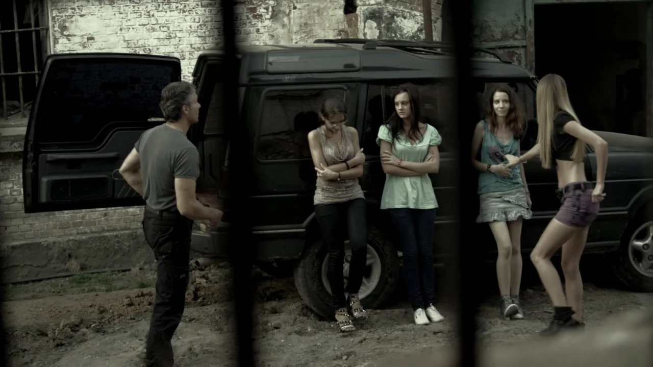 6 Bullets (2012) movie still: fullbody — A dusty outdoor lot features a dark SUV parked against a weathered brick wall. A group of five people stand…