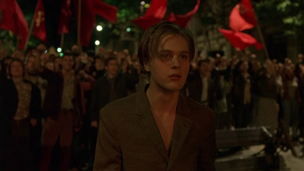 The Dreamers (2003) movie still: medium — A dense crowd holding red flags gathers on a city street at night. A young man stands in the foreground,…