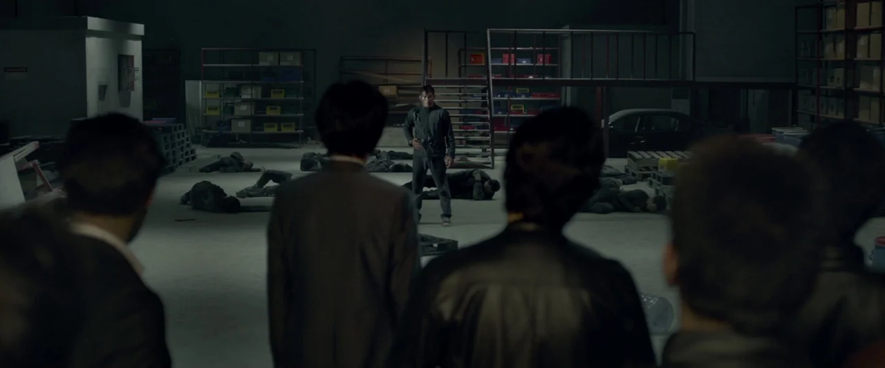 The Raid 2 (2014) movie still: wide — A spacious industrial storage room contains metal shelving units and scattered fallen bodies on concrete. A…
