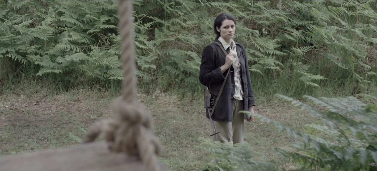 We Go In at Dawn (2020) movie still: medium — A woman stands in a lush woodland area with dense green ferns. She holds a rifle strap across her…