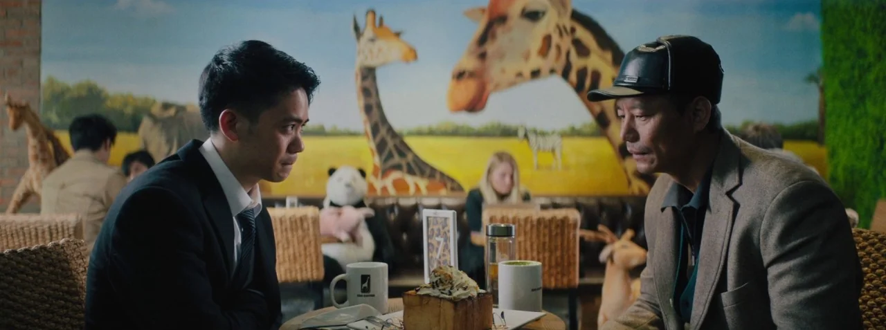 Dead Pigs (2018) movie still: medium — A cafe interior featuring a large safari-themed mural depicting giraffes and zebras. Two men sit at a small…