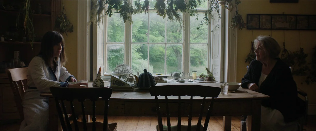 Fréwaka (2024) movie still: wide — A rustic dining room features a long wooden table set in front of a window. Two women sit at the table with dried…