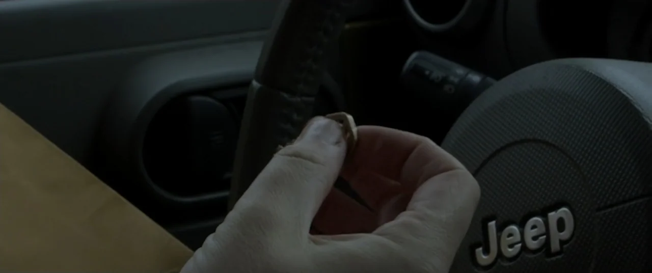 Amityville Cult (2021) movie still: closeup — The interior of a vehicle features a dark grey steering wheel with a visible logo. A hand holds a…