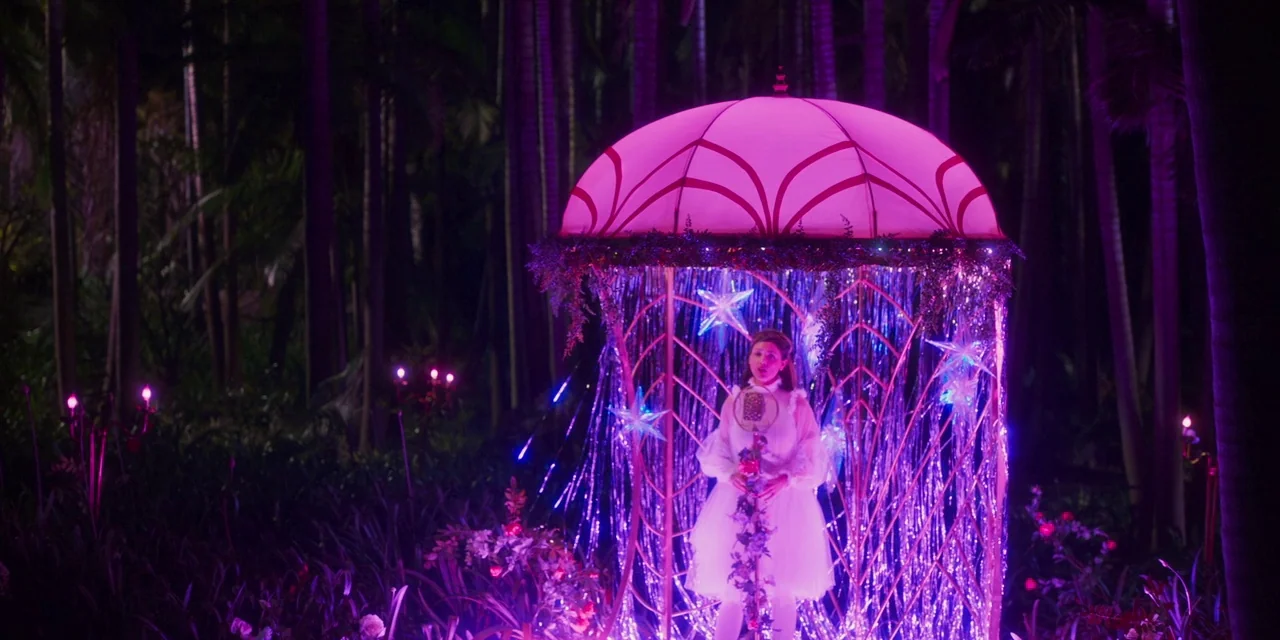 Paradise Hills (2019) movie still: medium — A woman stands within a jellyfish-shaped neon structure placed in a dark, wooded park area. She holds a…