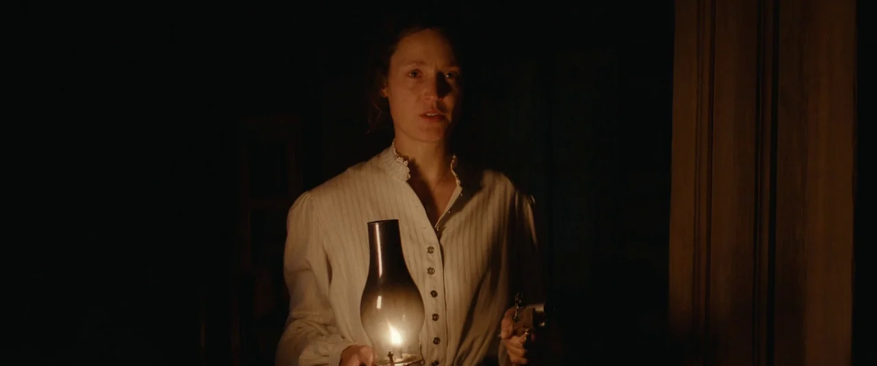 The Dead Don’t Hurt (2023) movie still: medium — A dimly lit interior space features a dark, unadorned room background. A woman holding an oil lamp…