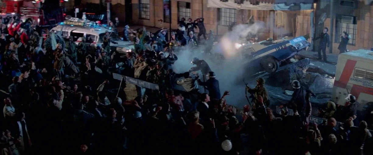 Ghostbusters (1984) movie still: wide — A dense urban intersection is filled with a large crowd and emergency response vehicles. A police car is…