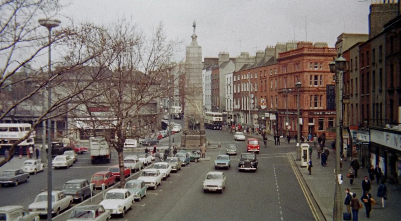 The Iguana with the Tongue of Fire (1971) movie still: wide — An urban intersection in 1960s Dublin features vintage cars and a central stone…