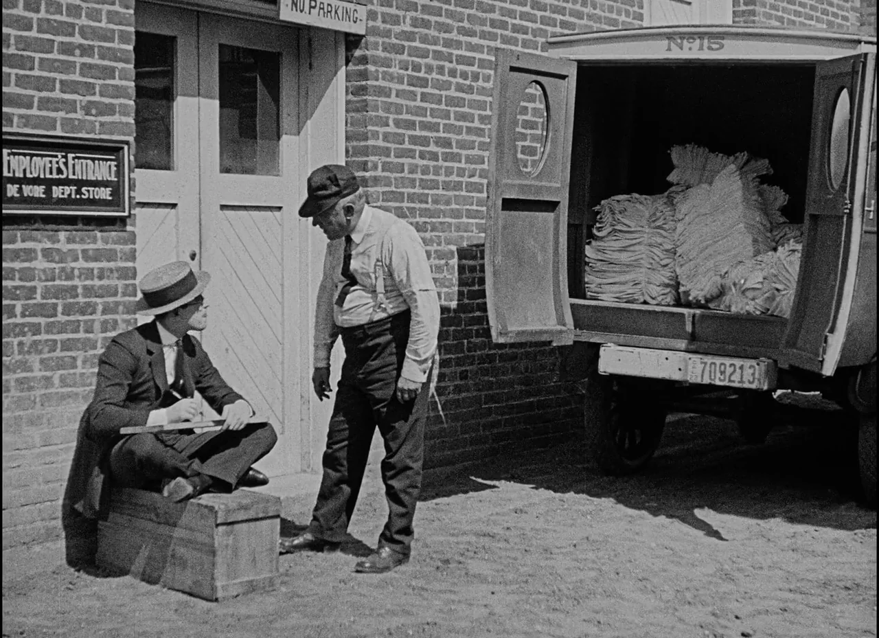 Safety Last! (1923) movie still: fullbody — An exterior brick wall with an entrance labeled for employees and a parked delivery van. A man sits on a…