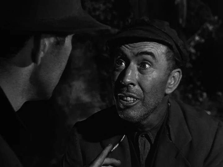 Nightmare Alley (1947) movie still: medium — A dark indoor space with minimal background detail visible in the shadows. A man in a cap speaks…