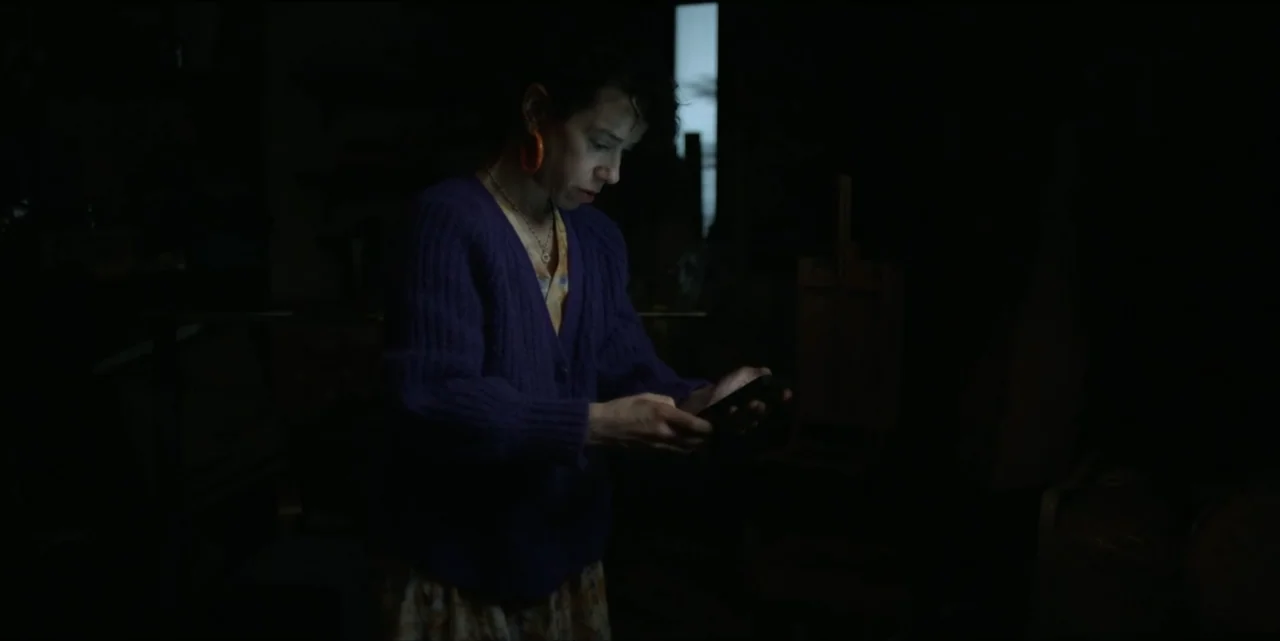 Bring Her Back (2025) movie still: medium — A dimly lit room contains art equipment in the dark background. A woman in a purple cardigan examines a…