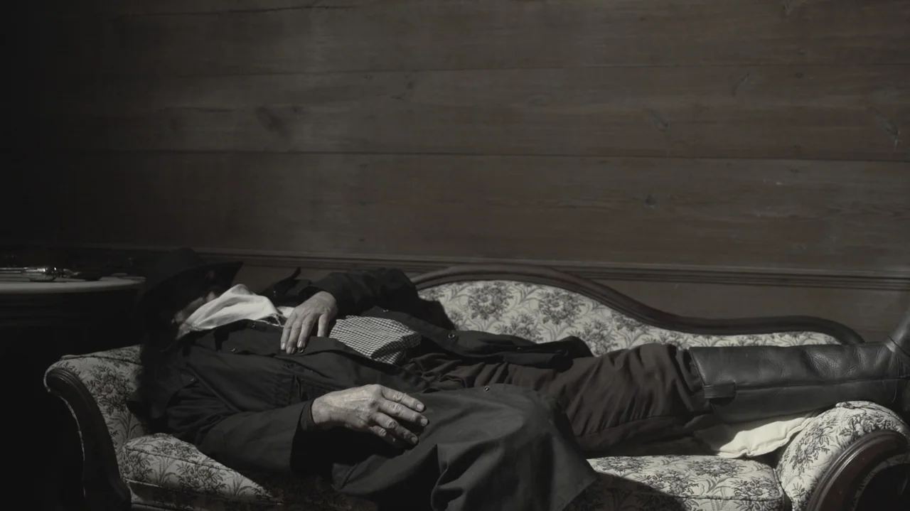 The Dalton Gang (2020) movie still: medium — A dimly lit room features wood-paneled walls and a patterned vintage fabric settee. A man in a duster…