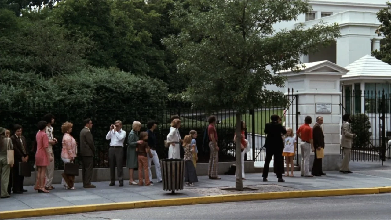 All the President's Men (1976) movie still: wide — A diverse crowd of people waits in a queue on a city sidewalk. The White House perimeter fence…