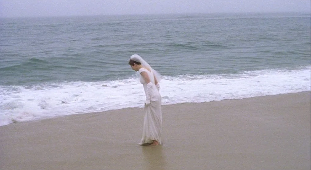Birth (2004) movie still: fullbody — A sandy shoreline under an overcast sky stretches across the entire frame. A woman in a white wedding gown…