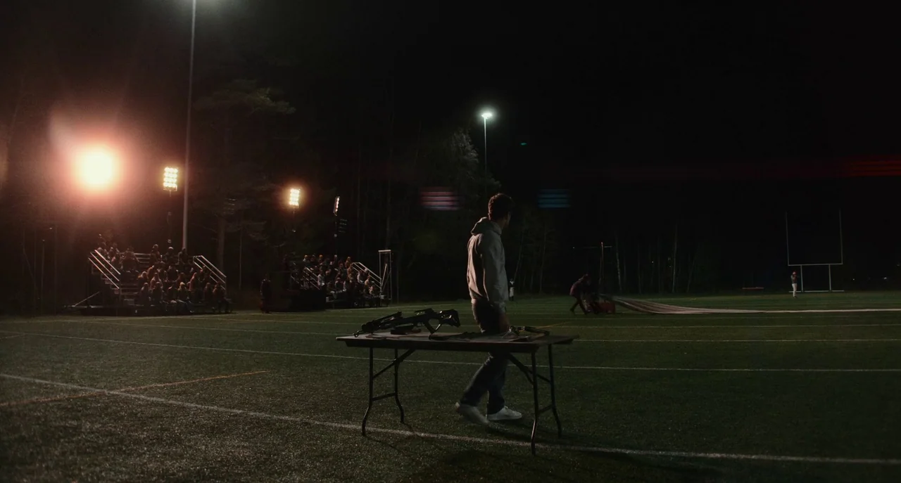 Dual (2022) movie still: wide — A night view of a football field illuminated by intense artificial stadium floodlights. A lone man stands beside a…