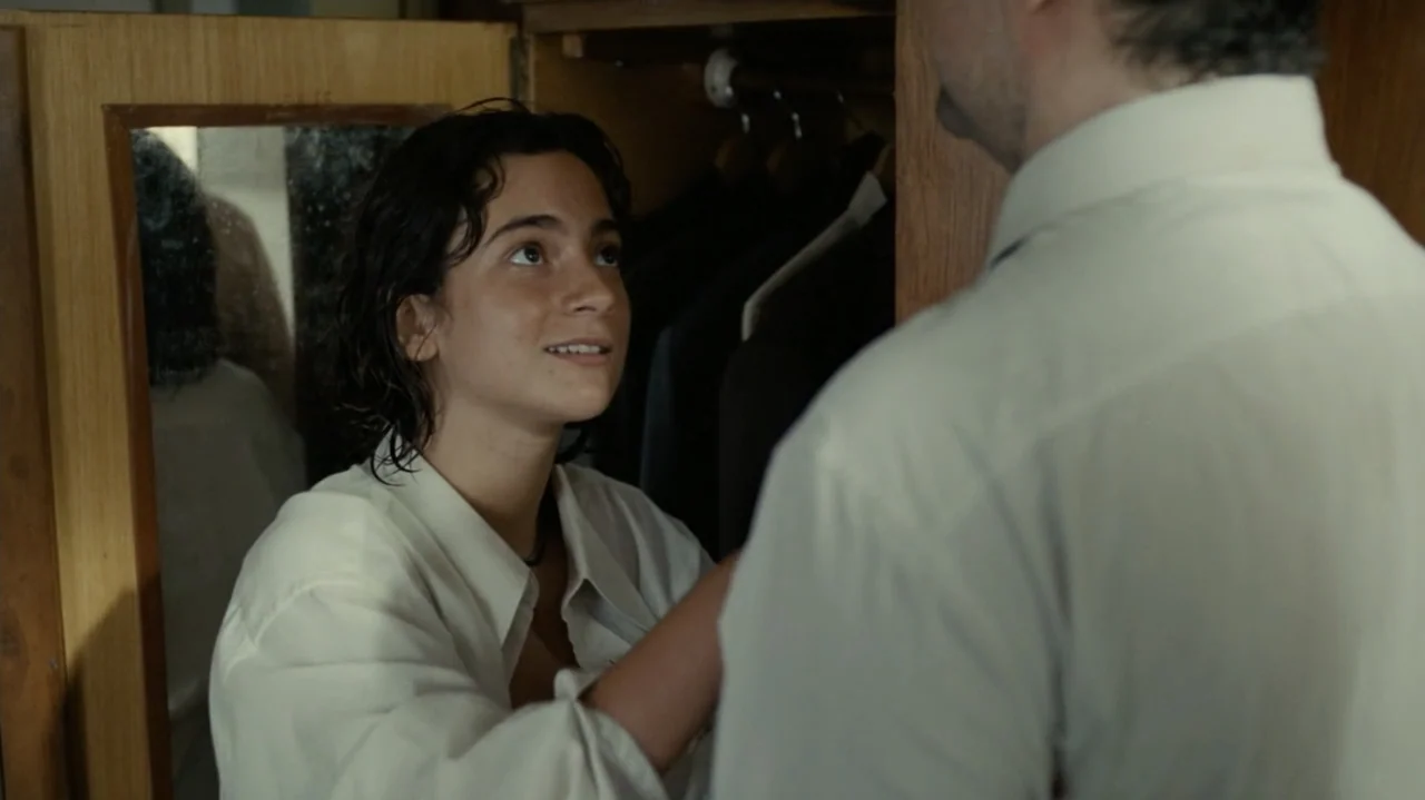 I'm Still Here (2024) movie still: medium — A walk-in closet filled with hanging garments inside a home. A woman looks up at a man standing before a…