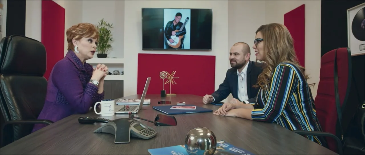 Loving is Losing (2019) movie still: medium — A modern office conference room features a dark wood table and red wall accents. Three people sit at…