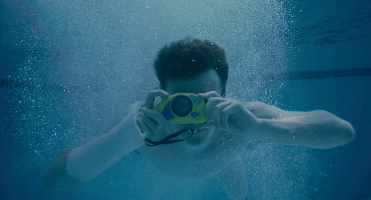 Aftersun (2022) movie still: medium — An underwater view of a pool with blue water and surrounding bubbles. A young man with brown hair holds a…