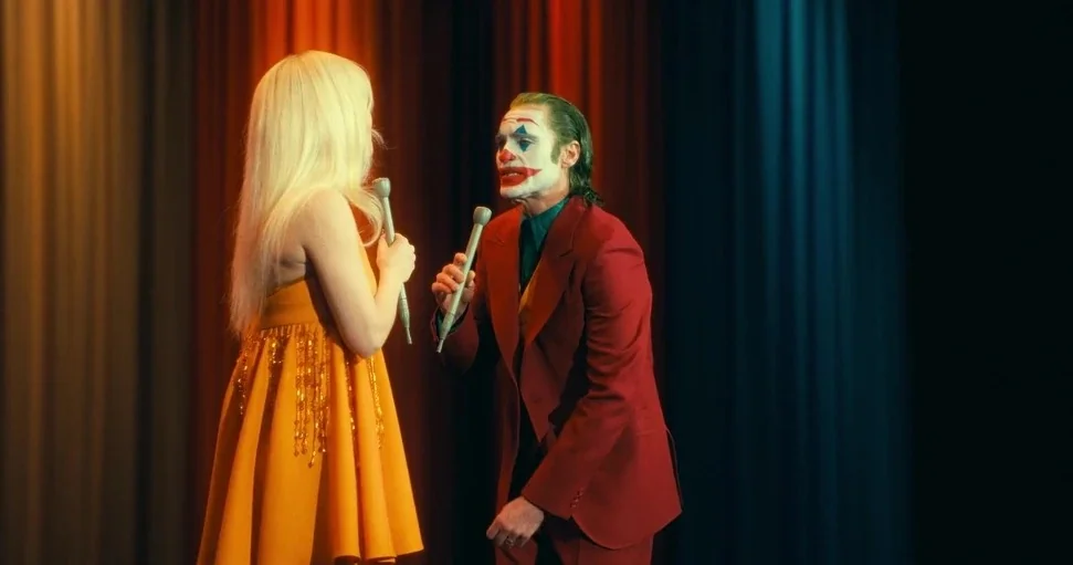Joker: Folie à Deux (2024) movie still: medium — A man in theatrical clown makeup and a woman stand on a television studio set. They each hold…