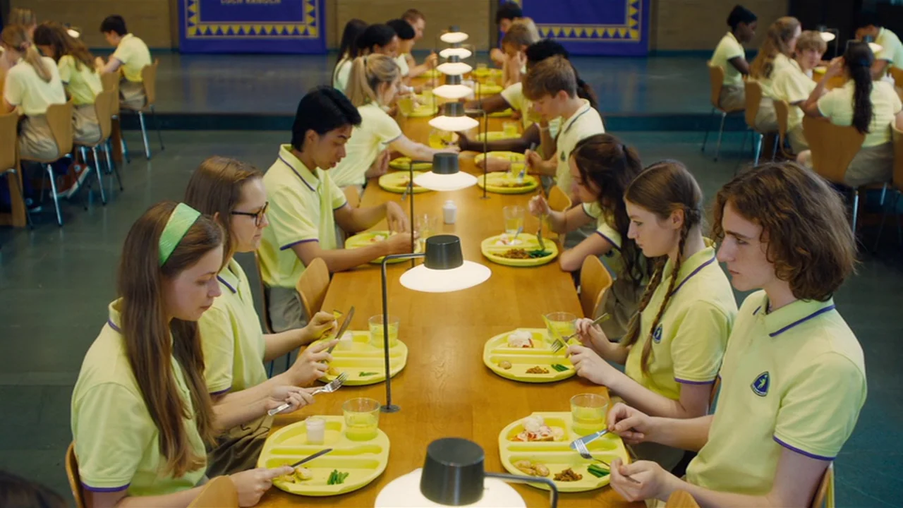 Club Zero (2023) movie still: medium — A long row of students in matching yellow shirts sit at wooden tables eating lunch. The school cafeteria is…