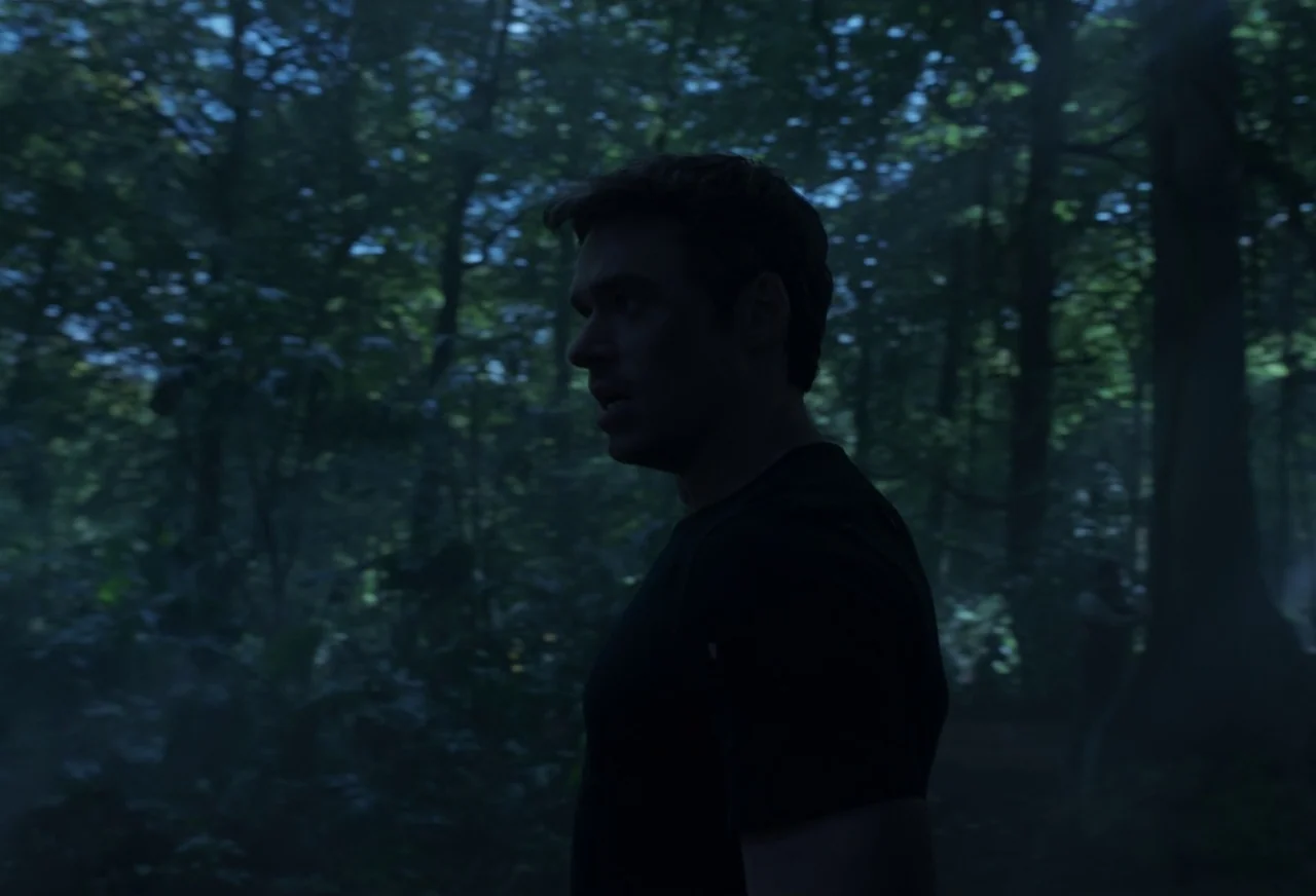 Eternals (2021) movie still: medium — A dense forest at night is shrouded in deep blue shadows and foliage. A man stands in the foreground, his…