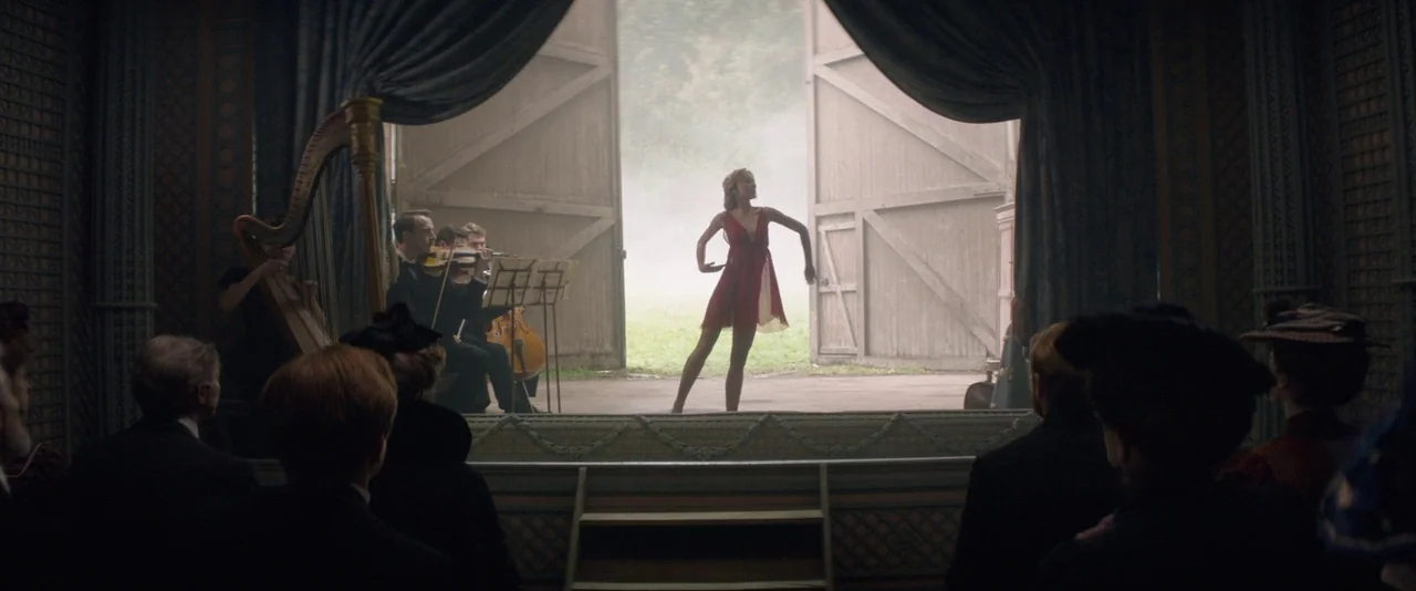 The Dancer movie still: wide — An interior space with wooden barn-style doors open to a misty outdoor field. A woman in a red dress dances for an…
