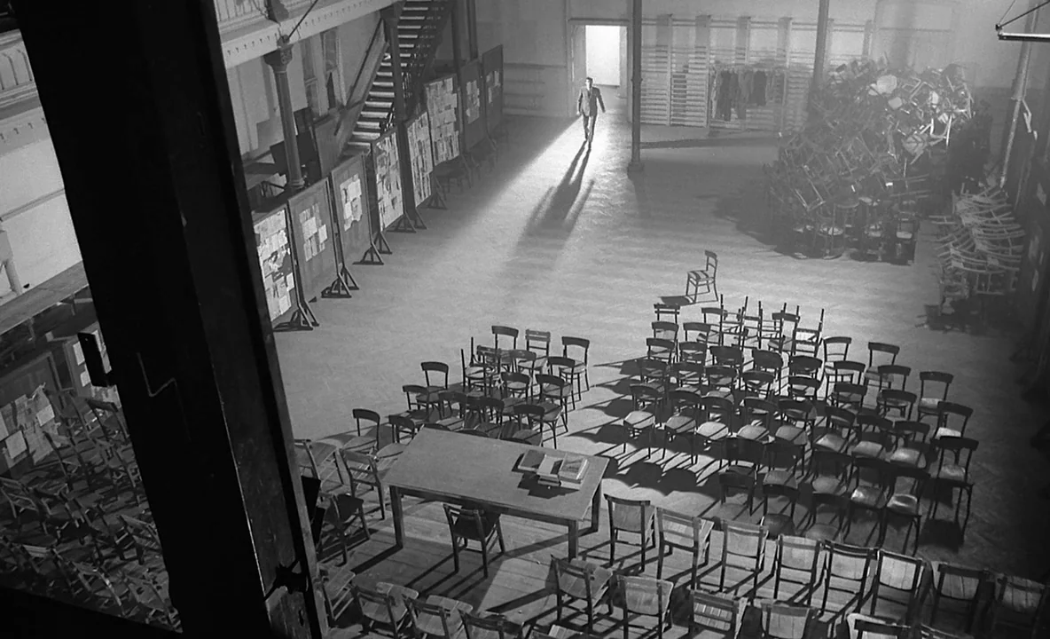 The Trial (1962) movie still: wide — A large, sparsely populated hall contains rows of empty wooden chairs and a table. A man enters through a…