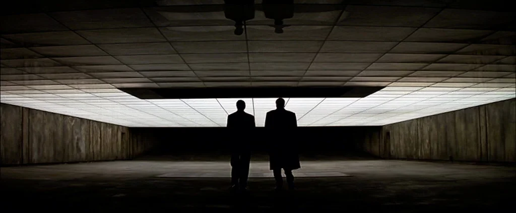The Dark Knight (2008) movie still: wide — A vast, minimalist concrete bunker space features a large luminous panel overhead. Two men in dark suits…