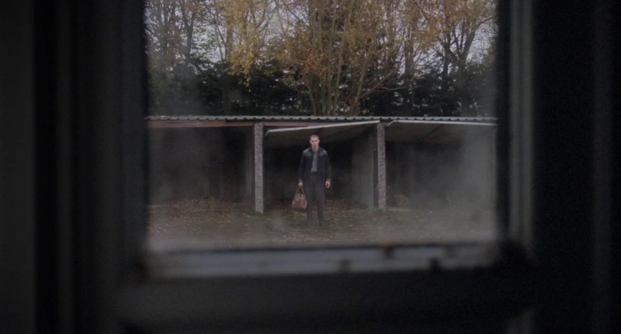Possum (2018) movie still: wide — A set of concrete lock-up garages stands beneath a line of autumnal trees. A young man wearing a dark jacket…