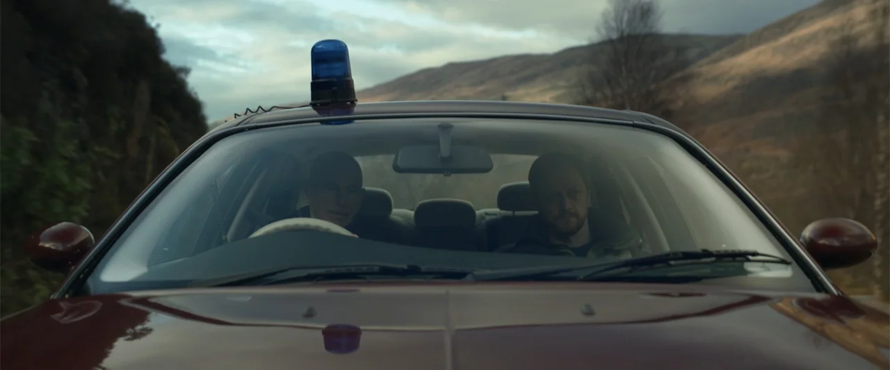 My Son (2021) movie still: medium — A maroon car with a blue light on the roof drives along a road. Two men sit inside the vehicle in front of a…