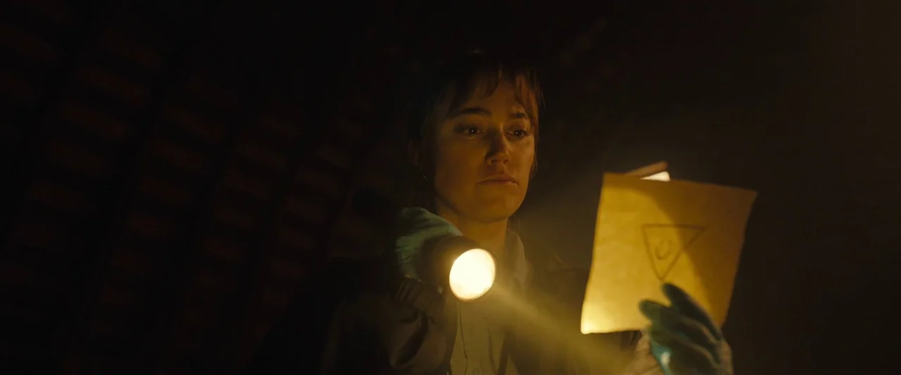 Longlegs (2024) movie still: closeup — A woman stands in a dark space lit only by her own light source. She examines a paper with a drawn triangle…