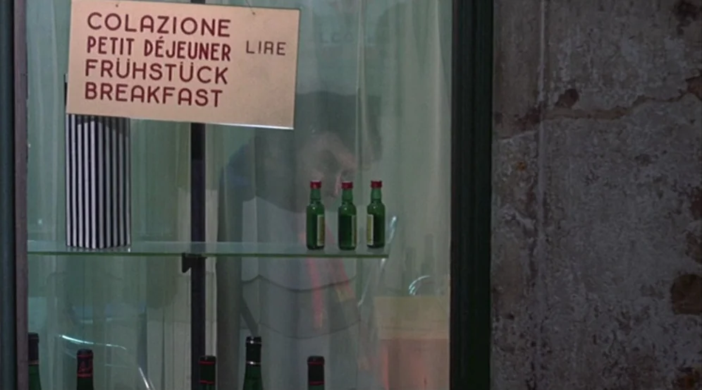 Don’t Look Now (1973) movie still: medium — A shop window displays small green bottles on glass shelves behind a multilingual sign. A man in a white…