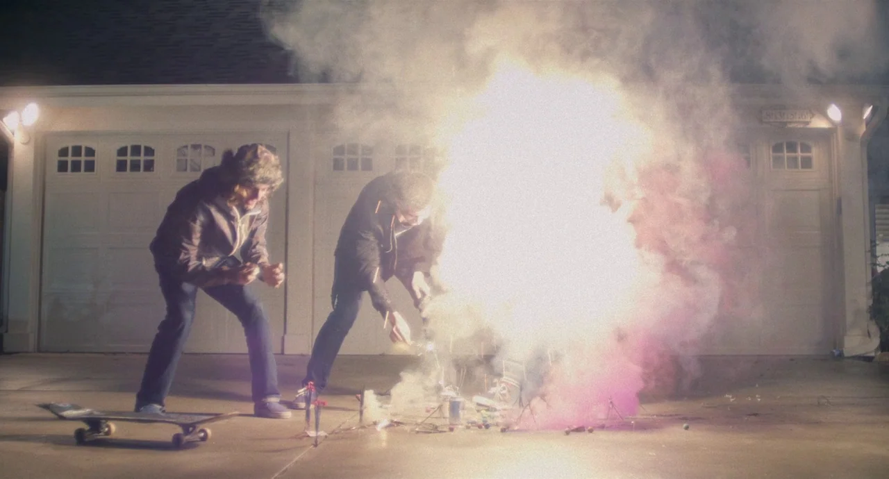 Palo Alto (2013) movie still: fullbody — A suburban residential driveway at night features two young men setting off fireworks. Bright light erupts…