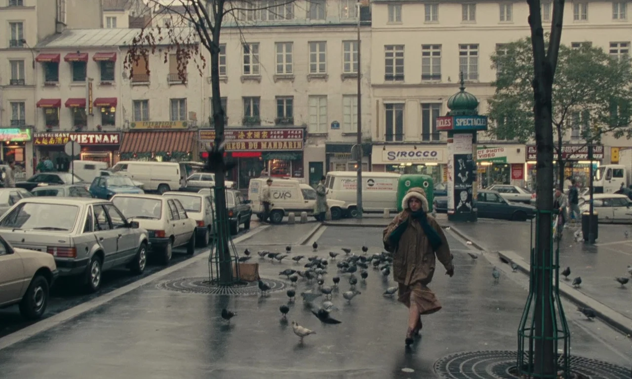 A Tale of Winter (1992) movie still: wide — A wet Paris city square features vintage cars, storefronts, and a Morris column. A woman in a parka…