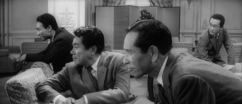 The Bad Sleep Well (1960) movie still: medium — Four men in suits sit on plush patterned couches in an opulent hotel lobby. They are oriented toward…