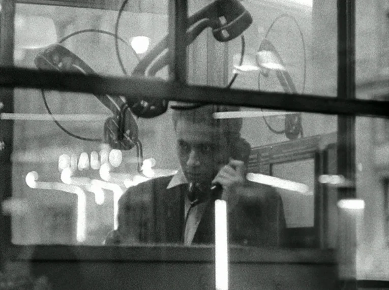 Le Petit Soldat (1963) movie still: medium — A man stands inside a glass enclosure at night, seen through reflections on the glass. A bright neon…