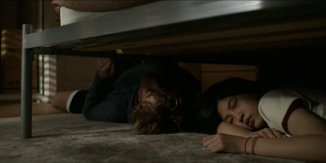 Bring Her Back (2025) movie still: medium — A carpeted bedroom floor beneath a metal bed frame is partially obscured by darkness. Two teenagers lie…