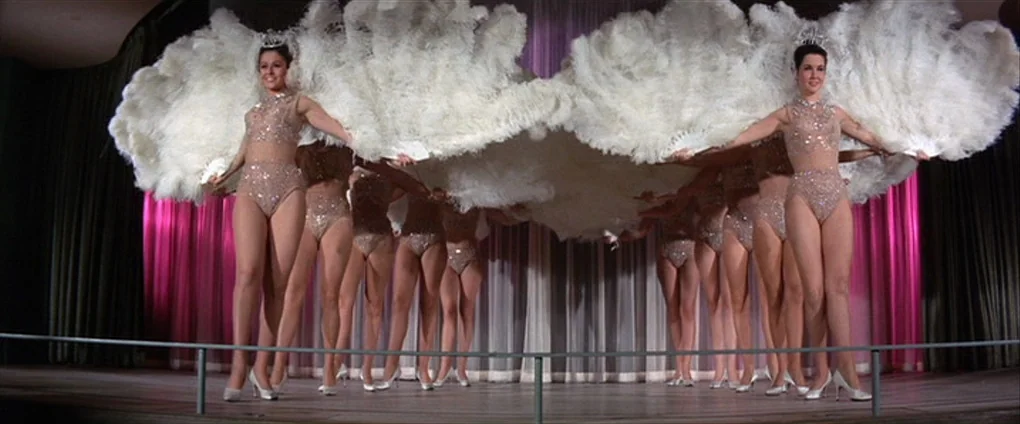 Viva Las Vegas (1964) movie still: fullbody — A group of showgirls stands on a wooden stage with vibrant pink back curtains. The performers are…