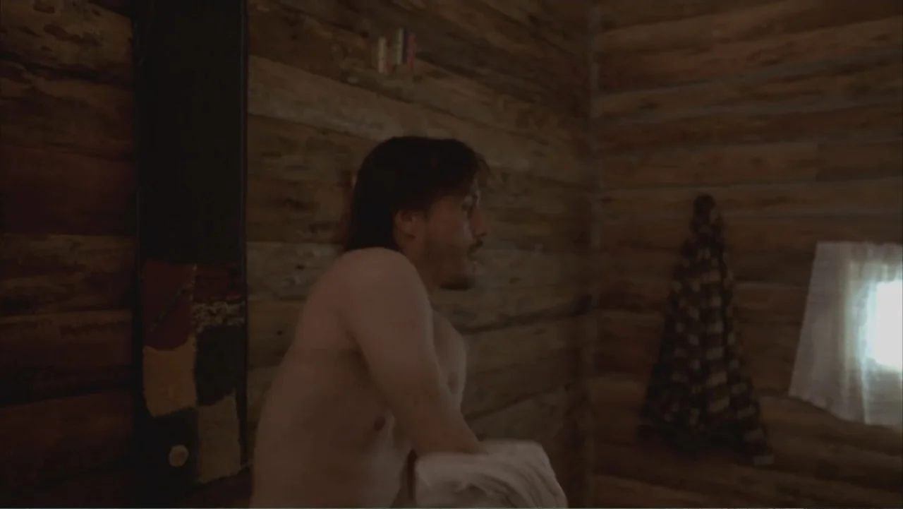 A Distant Place (2020) movie still: medium — A rustic cabin interior features walls constructed of horizontal wooden logs. A shirtless man sits in…