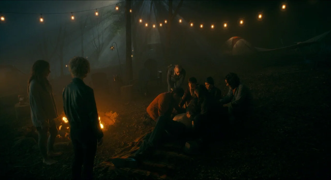 Doctor Sleep movie still: wide — A dark, fog-filled campsite at night with multiple people huddled around a small campfire. String lights hang…