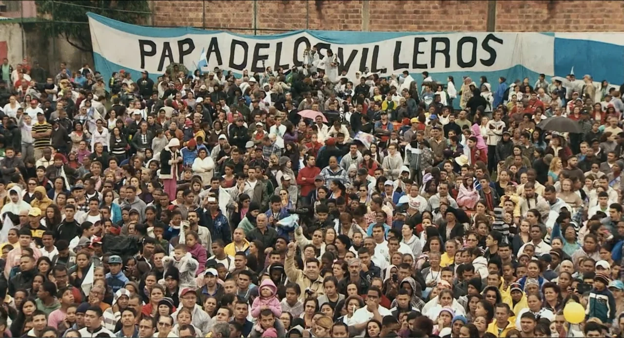 Pope Francis: A Man of His Word (2018) movie still: wide — A dense crowd of people gathers outdoors in front of a tall brick wall. A large blue and…