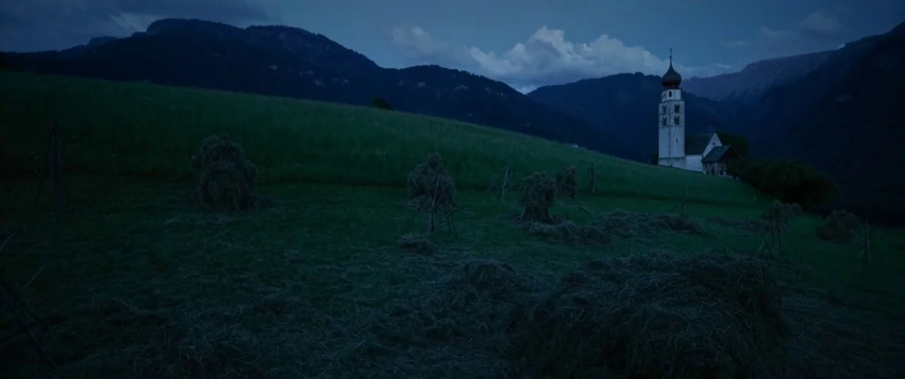 A Hidden Life (2019) movie still: wide — A rural hillside features scattered haystacks resting on a grassy meadow under dark mountains. In the…