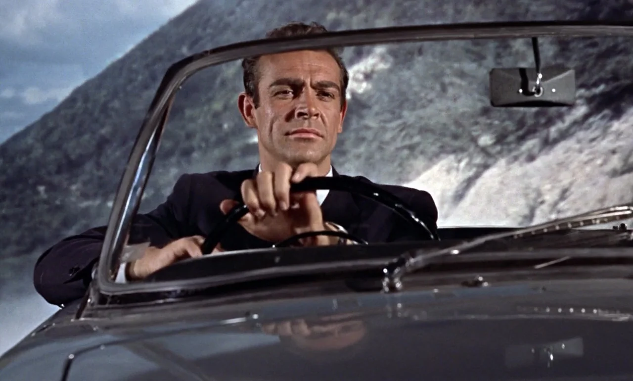 Dr. No (1962) movie still: medium — A man drives a convertible car along a winding mountain road. The steep rocky slope rises prominently in the…