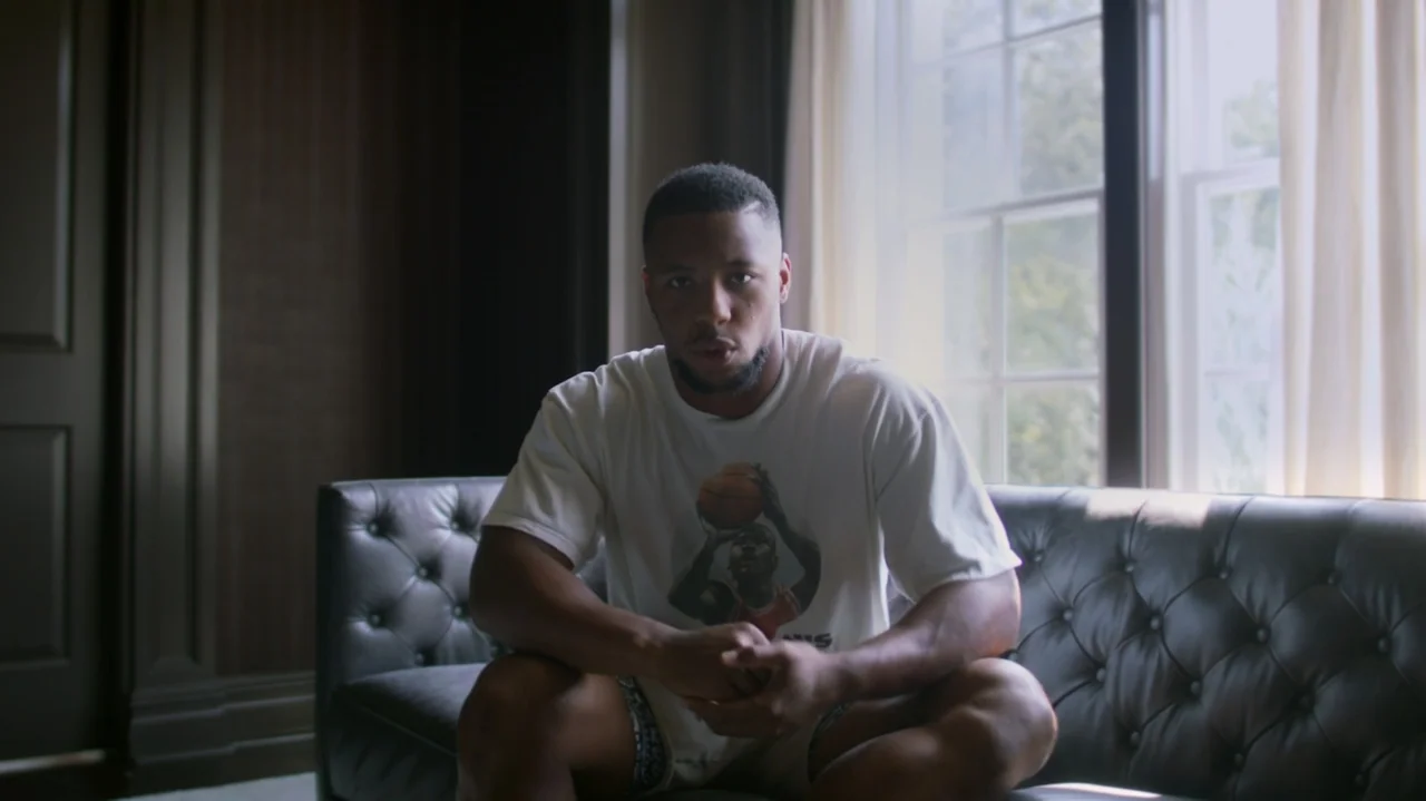 SAQUON (2025) movie still: medium — A man sits on a black leather couch in front of a bright window. The subject is wearing a graphic white t-shirt…