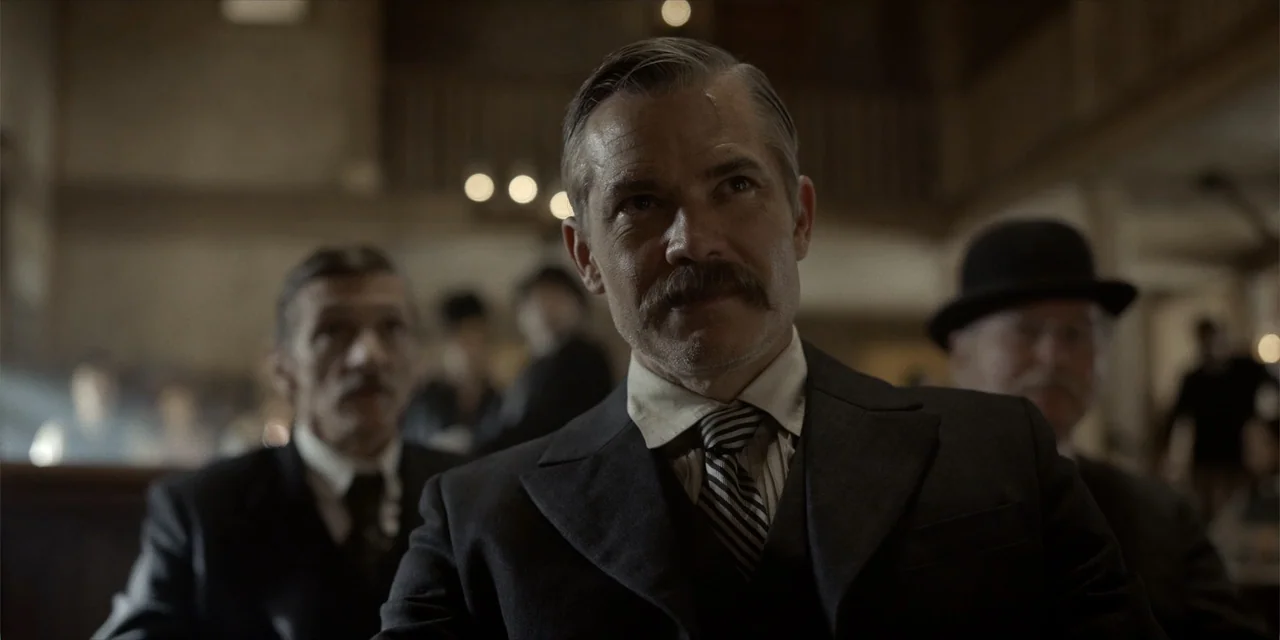 Deadwood: The Movie (2019) movie still: medium — A dimly lit room with wooden furnishings and a crowd of people in suits. A man sits in the…