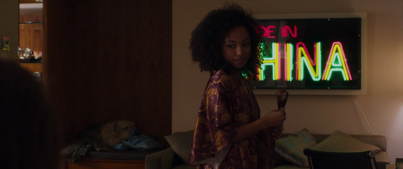 The Perfection (2018) movie still: medium — A hotel room features a dark wood panel wall and a neon sign display. A young woman in a paisley robe…